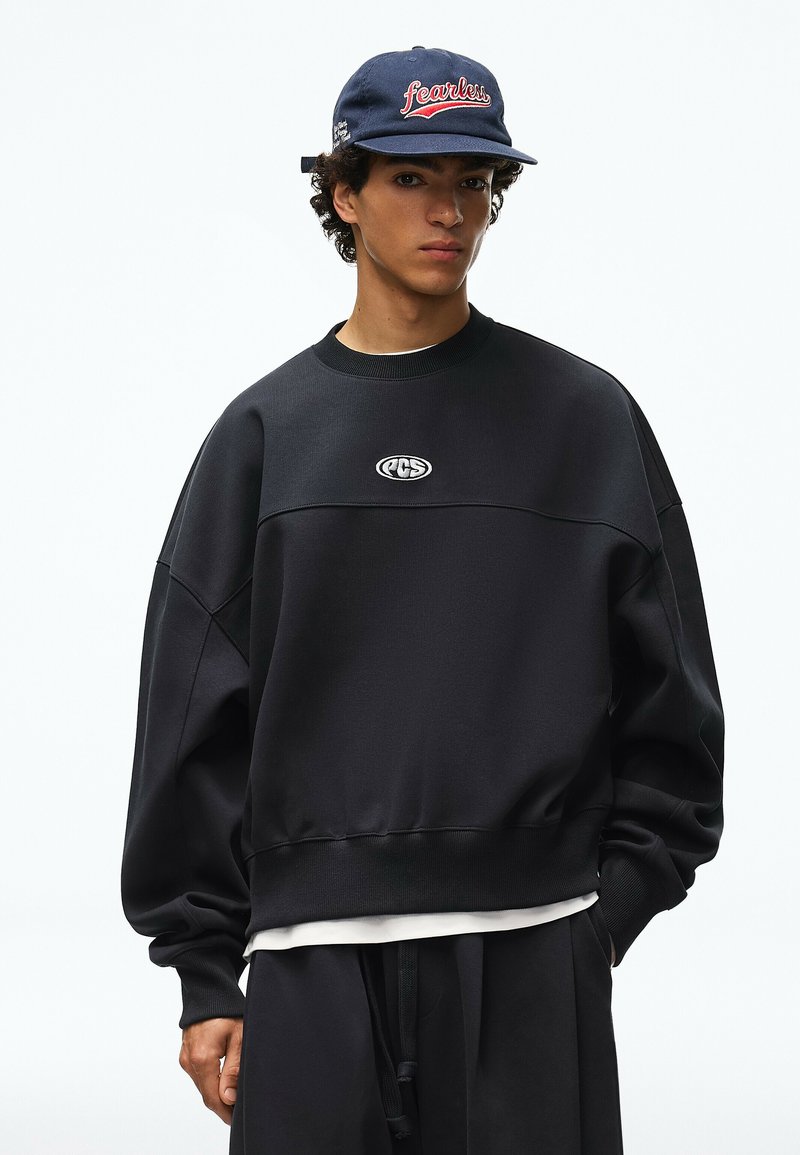 Jeune homme portant un sweat-shirt noir oversize avec un petit logo blanc et une casquette bleu marine avec un texte rouge "fearless".