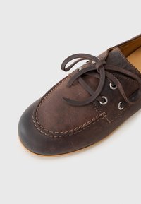 Sapatilha de barco em couro castanho com bico arredondado, sola de borracha escura contrastante e design de atacadores angulado com ilhós prateados e detalhes de costura.