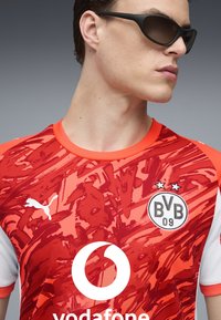 Maglia sportiva rossa con motivi astratti, dettagli arancioni e maniche bianche. Presenta il logo Puma e l'emblema Vodafone. Due stelle sopra il distintivo del club.