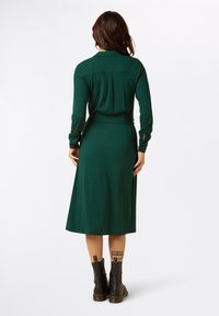 Grünes, gepunktetes, langärmliges Kleid mit tailliertem Schnitt und ausgestellt Rock. Verfügt über einen Kragen und Knöpfe an den Ärmeln. Getragen mit schwarzen Stiefeln.