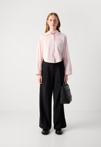 Camicia corta button-up rosa chiaro, pantaloni larghi neri, borsa nera con motivo geometrico e scarpe nere. Il modello è in piedi davanti a uno sfondo semplice.