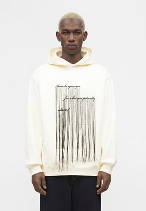 Mladi moški s kratkimi svetlo blond lasmi nosi kremasto kapucar z črnimi navpičnimi črtami, ki spominjajo na vrvice, in napisom spredaj.