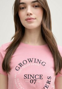 Camiseta de algodón de color rosa claro con un diseño gráfico circular en negro. El texto incluye "GROWING," "SINCE 07" y "TOM TAILOR DENIM." Mangas cortas.
