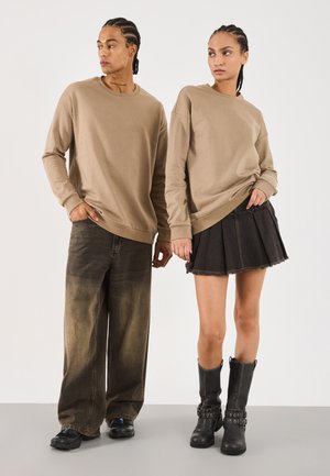 Zwei Personen mit geflochtenem Haar, die passende beige Sweatshirts tragen; eine in weit geschnittenen, ausgewaschenen Jeans und Turnschuhen, die andere in einem schwarzen Rock und schwarzen Stiefeln.