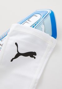 Hvide sportsskinner med sort Puma-logo og blå beskyttende foddækning delvist synlig indeni.