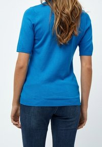 Kortärmad tröja i klart blått stickat material, med ribbat halsringning och fåll, tillsammans med mörkblå jeans.