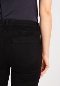 Svarta högmidjade jeans med en slät yta, med en bakficka och bälteshällor, tillsammans med en enkel mörk topp.