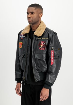 Hombre con chaqueta bomber de cuero negro con cuello de borreguillo color marrón claro y múltiples parches de aviación de colores, de pie frente a un fondo blanco.