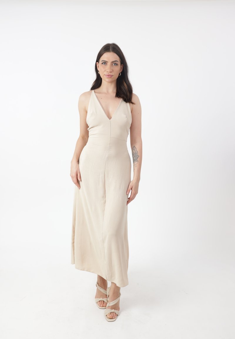 Beiges Maxikleid mit tiefem V-Ausschnitt, figurbetontem Oberteil und schwingendem Rock. Glatter, leichter Stoff mit einer dezenten Textur, kombiniert mit Riemchen-Pumps.