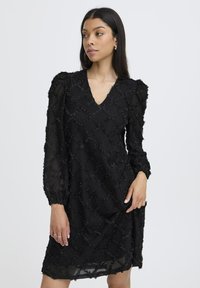 Sort kjole med V-hals, lange pufærmer og tekstureret stof med en diamantmønster og hævede detaljer. Midtlængde design.