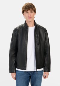 Zwarte leren jas met een staande kraag, ritssluiting aan de voorkant en gewatteerde schouderaccenten. Heeft een voorkantzak en een gladde afwerking. Draagt over een wit shirt.
