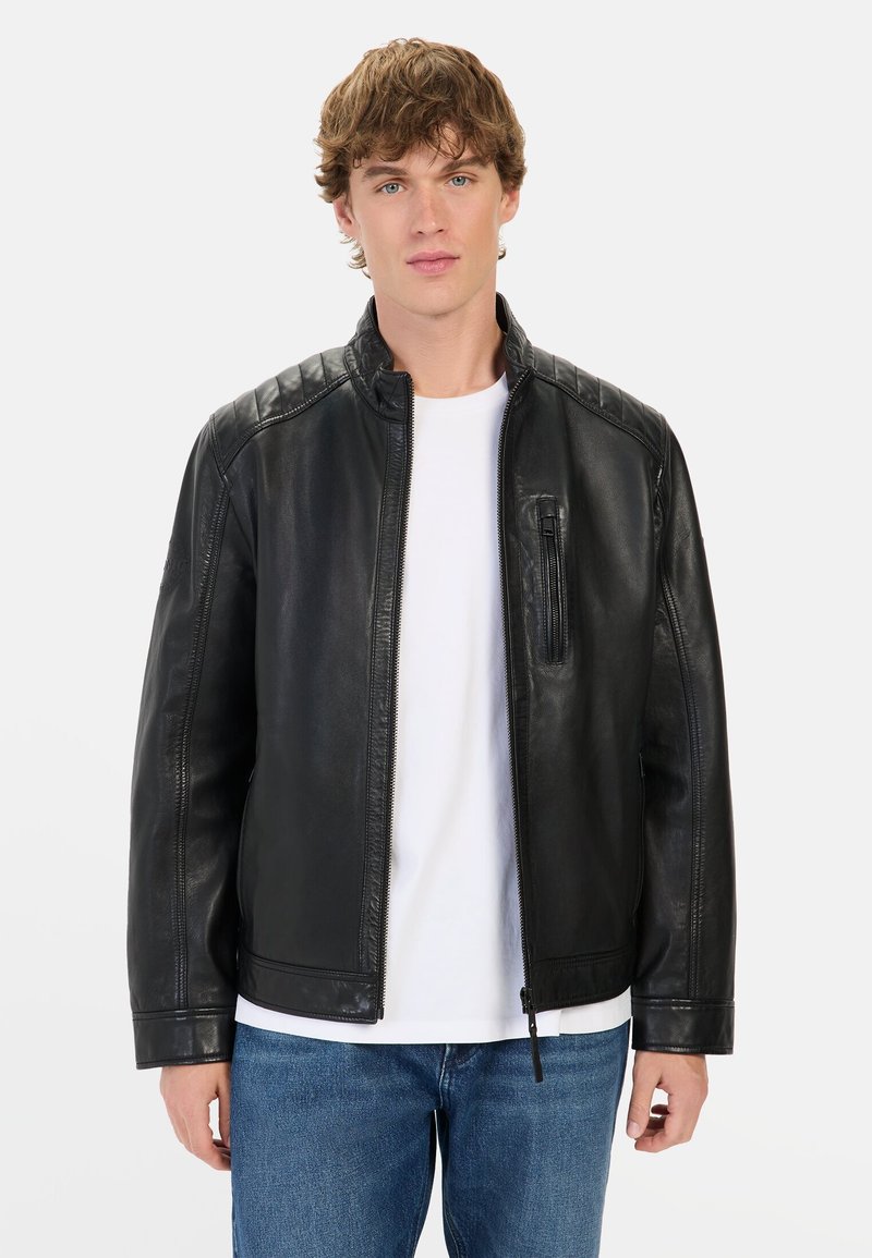 Zwarte leren jas met een staande kraag, ritssluiting aan de voorkant en gewatteerde schouderaccenten. Heeft een voorkantzak en een gladde afwerking. Draagt over een wit shirt.