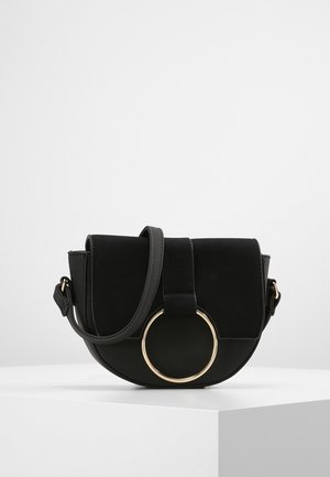 Zwarte halfronde crossbodytas met klep, verstelbare band en grote decoratieve gouden ring aan de voorkant, tentoongesteld op een witte ondergrond.