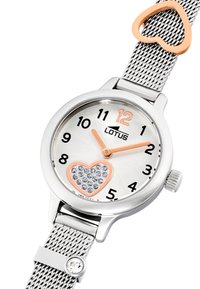 Reloj con correa de malla plateada, esfera redonda, diseño de corazón adornado con cristales, acentos en oro rosa y marcadores numéricos negros sobre un fondo plateado texturizado.