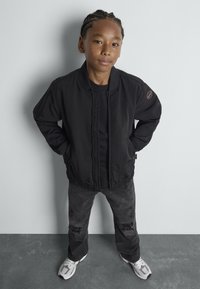Veste bomber noire avec des poignets et un col à côtes, portée par-dessus un T-shirt noir. Associée à un jean noir usé et des baskets blanches.