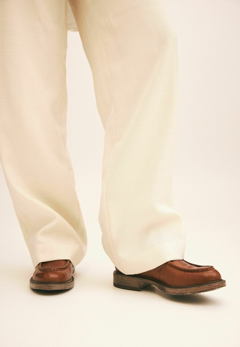 Personne portant un pantalon blanc ample et des chaussures en cuir marron, debout sur un sol clair devant un fond uni.