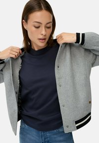 Grau wollener Bomberjacke mit schwarz-weißen gestreiften Bündchen, Druckknöpfen und einem gemusterten Innenfutter, getragen über einem marineblauen T-Shirt.