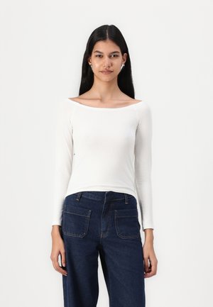 Hvid langærmet skjorte med off-the-shoulder halsudskæring. Kombineret med mørkeblå denim bukser med bred ben, der har to baglommer.