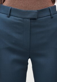 Προσαρμοσμένο σκούρο μπλε παντελόνι με κρυφό μπροστινό κούμπωμα, θηλιές ζώνης και ορατή υφασμάτινη καφέ μέση πλεκτού που είναι μέσα στο παντελόνι.