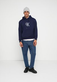 Marinblå luvtröja med framficka och dragsko. Matchas med ljusblå jeans och svarta skor. Grå stickad mössa.