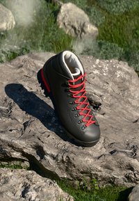 Schwarzer Wanderstiefel mit roten Schnürsenkeln, der auf einem großen grauen Stein liegt, umgeben von Gras und kleineren Steinen im Freien.