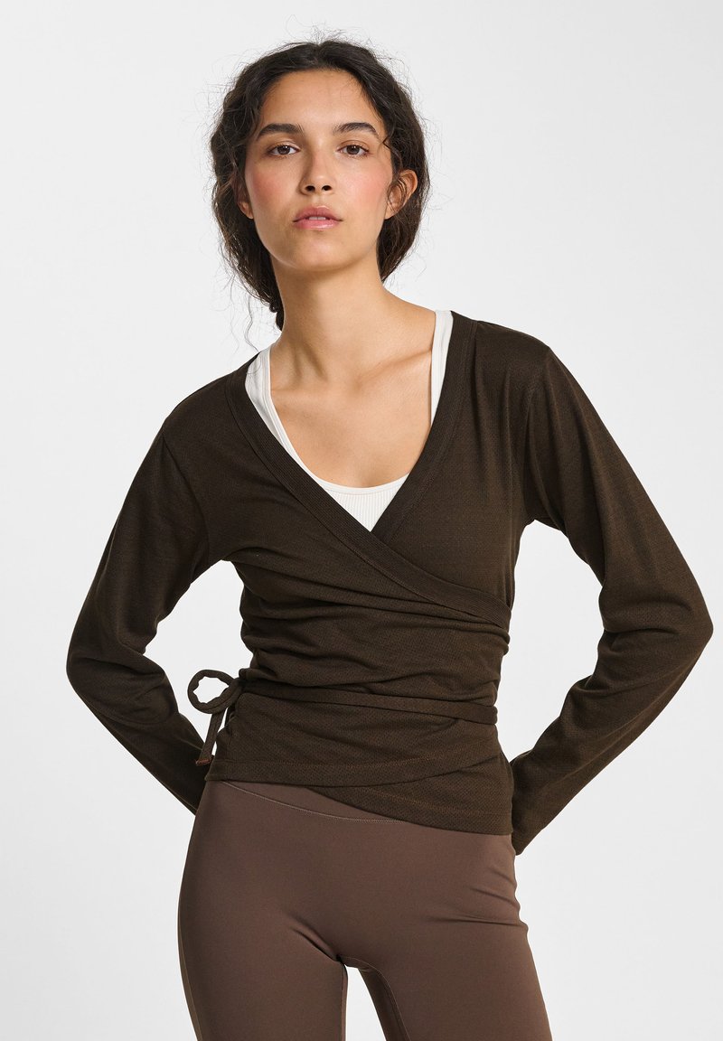 Bruine wikkeltop met lange mouwen en een strik in de taille, gelaagd over een witte tanktop. Glad textuur met een subtiele glans.