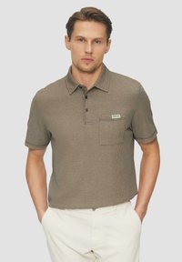 Kurzärmliges Poloshirt aus strukturiertem braunem Stoff mit Kragen, drei Knöpfen, einer Brusttasche und einem kleinen Etikett auf der Tasche.