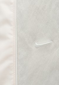 Ύφασμα ανοιχτού γκρι με κεντημένο λευκό λογότυπο Nike Swoosh, δίπλα σε πάνελ υφάσματος σε ανοιχτό ροζ με ραφές.