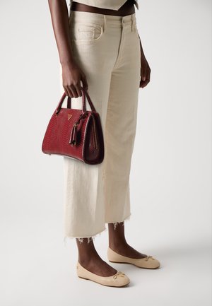 Sac à main texturé rouge avec une fermeture éclair et des pompons, tenu à la main. Pantalons courts de couleur claire avec des ourlets effilochés et des ballerines beiges.