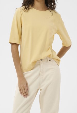 Mujer que lleva una blusa de manga corta de color amarillo pálido y pantalones de talle alto de color blanco roto, con una mano en el bolsillo y la otra descansando a su lado.