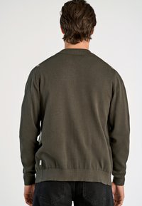 Donkergroen gebreid sweater met een ronde kraag, geribde manchetten en zoom. Heeft subtiele details op de schouders en een klein merklabel aan de zijkant.