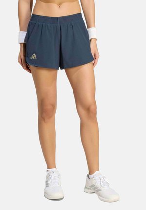 Frau trägt dunkelblaue Sportshorts, weiße Handgelenkbandagen und weiße Sportschuhe, steht vor einem schlichten Hintergrund.