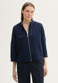 Veste hoodie zippée bleu marine avec capuche à cordon, manches 3/4 et tissu texturé. Zip en métal argenté et coupe décontractée.