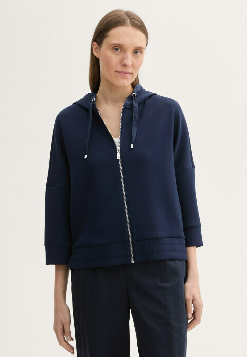 Veste hoodie zippée bleu marine avec capuche à cordon, manches 3/4 et tissu texturé. Zip en métal argenté et coupe décontractée.
