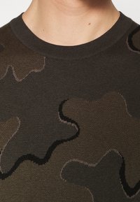 Strickpullover mit einem Camouflage-Muster in Schattierungen von dunklem Grün und Braun, mit kontrastierenden schwarzen Akzenten entlang der Struktur des Designs.