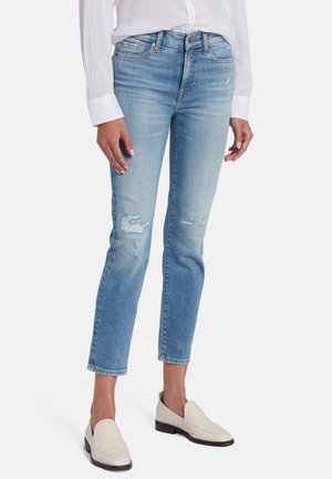 Hellblaue, ausgebleichte Jeans aus Denim. Sie haben eine schmale Passform, sind knöchellang mit Saumaufschlägen und zeigen sichtbare Abnutzungserscheinungen in der Kniegegend. Kombiniert mit weißen Slippern.