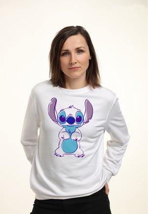 Kvinde med brunt hår iført en hvid sweatshirt med en stor, farverig tegneseriefigur med store øjne og store ører.