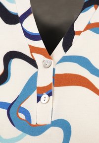 Vaaleanbeige lyhythihainen paita, jossa on monivärisiä aalto-kuvioita sinisen, oranssin ja tummansinisen sävyissä. Siinä on V-kaula-aukko ja kaksi valkoista nappia.