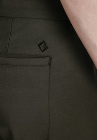 Nærbillede af et mørkebrunt bukselomme med syede sømme og et lille sort logo-patch over lommen, ved siden af en persons arm.