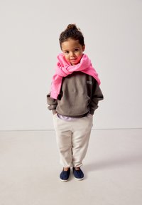 Kind mit lockigem Haar in braunem Sweatshirt und cremefarbener Hose, rosa Pullover über den Schultern, steht mit den Händen in den Taschen und lächelt.