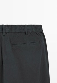 Schwarze Hose mit elastischem Bund, zwei Gürtelschlaufen und einer unauffälligen Gesäßtasche mit horizontaler Öffnung. Glatte, matte Textur.