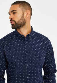 Navy-blaues Hemd mit weißem Diamantmuster. Verfügt über einen klassischen Kragen, lange Ärmel und eine durchgeknöpfte Vorderseite, gefertigt aus einem glatten Stoff.