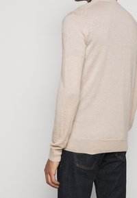 Pull léger beige à manches longues et encolure ronde. Le tissu semble lisse avec une texture subtile. Porté avec un jean foncé.