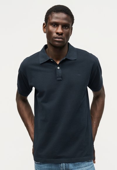 Νέος άνδρας που φοράει σκούρο μπλε πόλο με κοντά μανίκια και ανοιχτόχρωμο τζιν, στέκεται με τα χέρια πίσω από την πλάτη σε απλό φόντο.