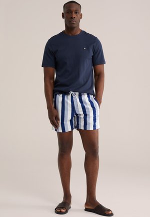 Muž v námornícky modrom tričku, modrých a bielo pruhovaných plavkových šortkách a tmavých šlapkách stojaci s jednou rukou v vrecku.