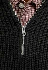 Silberner Reißverschluss, der teilweise an einem schwarzen gestrickten Pullover geöffnet ist, und ein darüber liegendes karminrot-weißes kariertes Hemd mit weißem Knopf darunter zeigt.