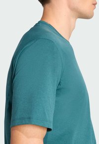 Camiseta de manga corta en color verde azulado, confeccionada con un tejido suave y texturizado. Presenta un cuello redondo y un corte holgado, con detalles de costuras visibles en las mangas.