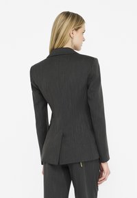 Blazer grigio scuro su misura con righe verticali, spalle strutturate e uno spacco singolo sul retro. Il tessuto appare liscio e testurizzato.