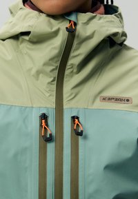 Giacca impermeabile di colore verde e teal. Presenta zip e dettagli a contrasto in arancione. Collo alto con cappuccio regolabile.