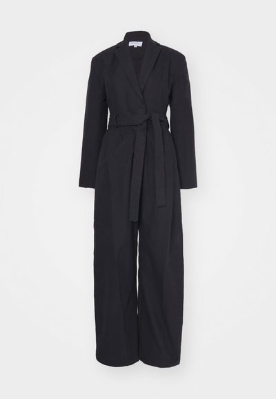 Salopetă neagră cu un design înfășurat, mâneci lungi și o curea în talie. Fabricată dintr-un material texturat cu o croială ajustată.